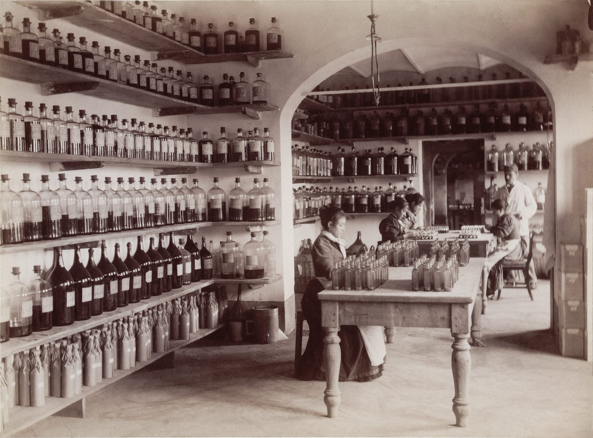 Operai, molte donne fra loro, tra scaffali di oli essenziali, assolute e distillati nello stabilimento di Corso Loreto, Milano, 1898